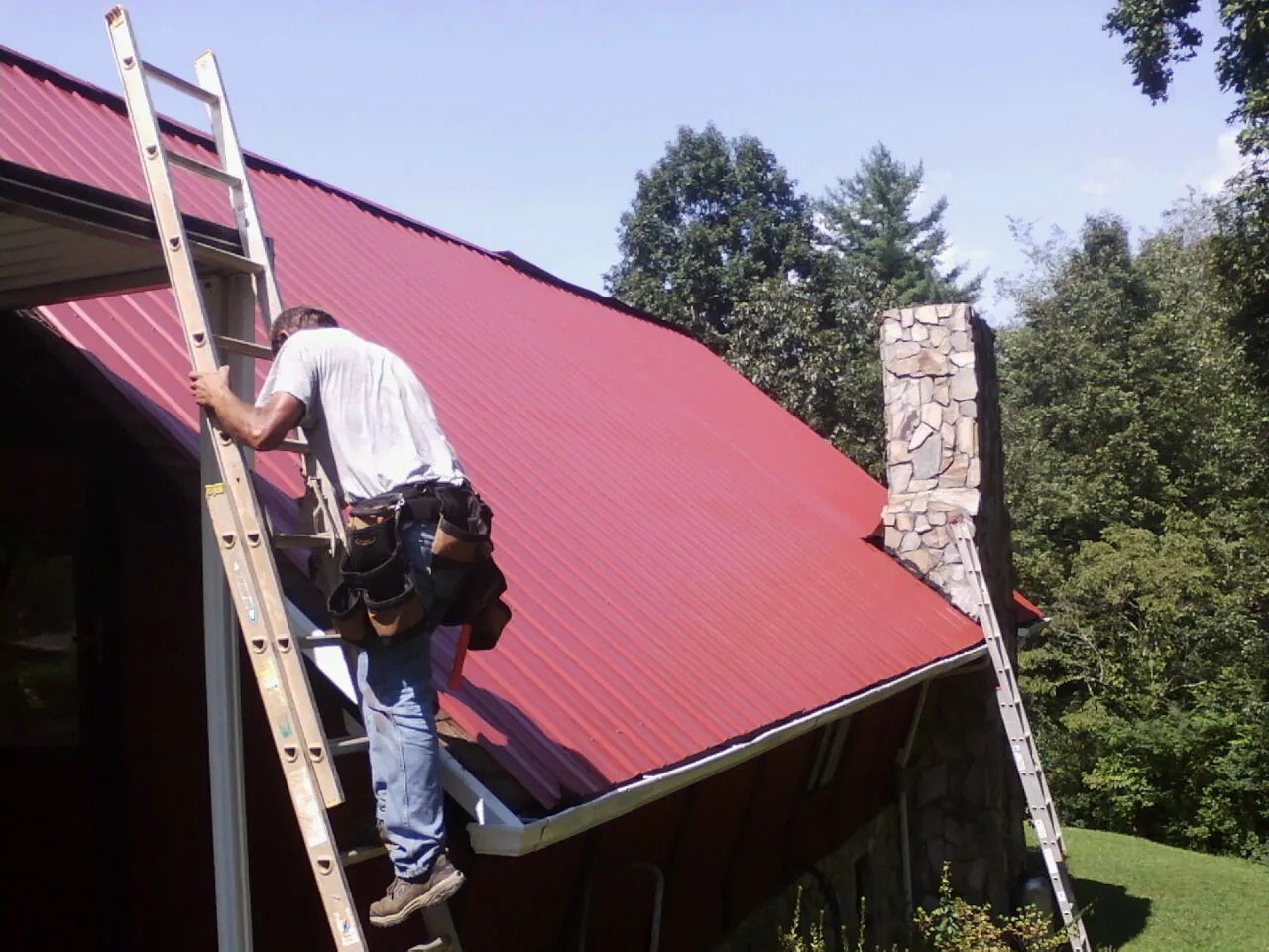 Roofer on a ladder beside a red metal roof and stone chimney — Soffit & Fascia Repair by Roofing Brothers in Canfield